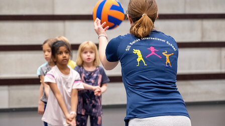 A sports teacher throws a volleyball to a child. You can only see her shoulders and her pigtail from behind. 