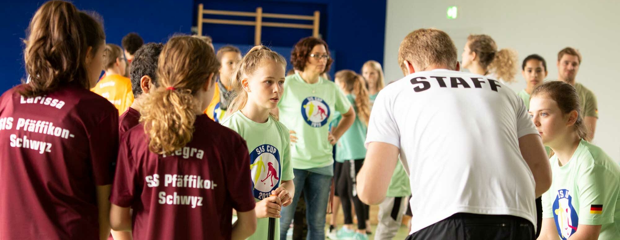 Eine Szene in einer Turnhalle. Ein Sportlehrer mit einem weissen T-Shirt, das "Staff" zeigt, steht vor einer Gruppe Schülerinnen und Schüler.