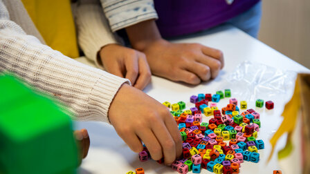 Nahaufnahme von drei Kinder-Händen auf einem Tisch. Eine Hand greift in einen kleinen Haufen bunter Buchstaben-Perlen.