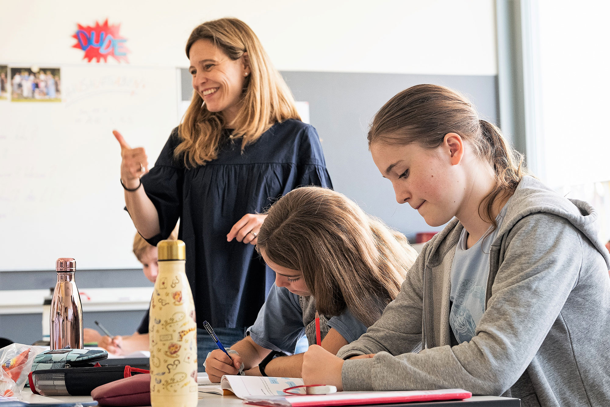 Zwei Mädchen schreiben etwas in ihr Heft und im Hintergrund steht die Lehrerin lachend und fröhlich. 	