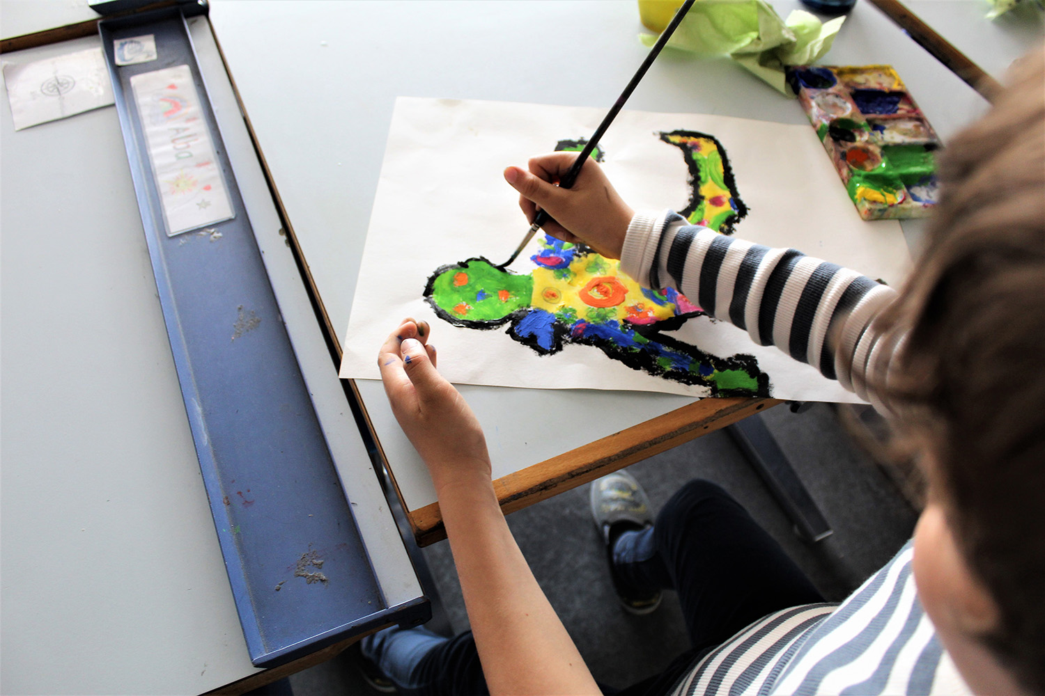 A child's hand paints a colourful little man with a brush on paper on the desk. You can see the back of the child's head, the painting hand and the motif.