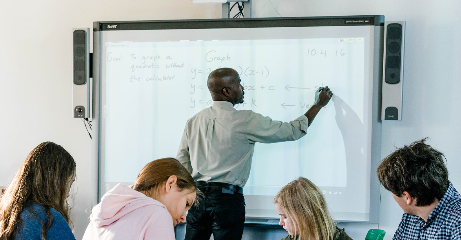 In a mathematics lesson, a teacher illustrates an issue on a smartboard. He is seen from the side at an angle.