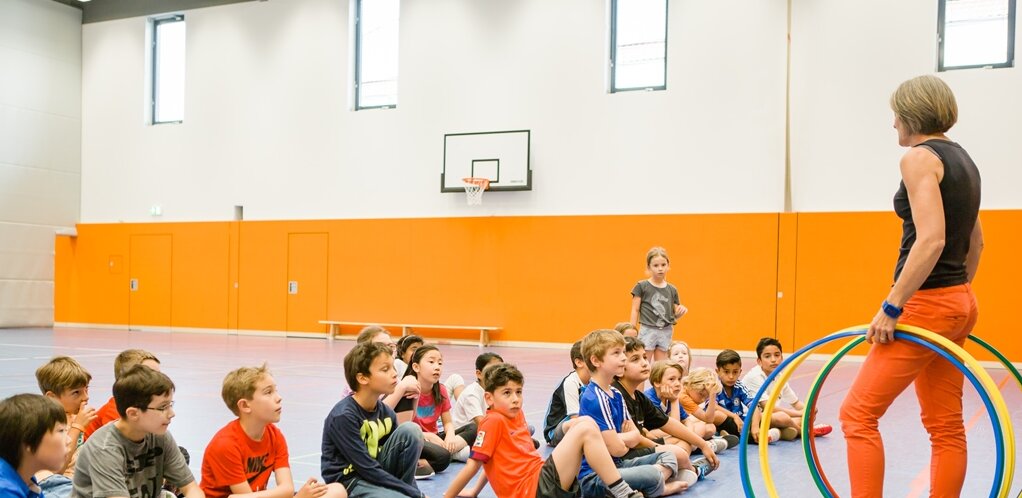 In a sports hall, a group of students sit on the floor and look at the standing teacher who is holding several hula hoops in her hand. In the background you can see an orange wall.