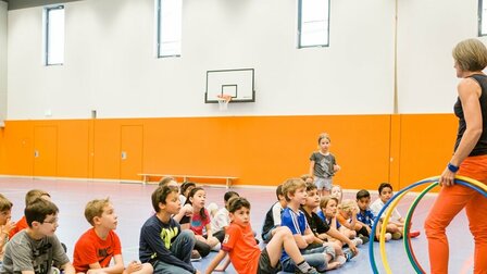 In einer Turnhalle sitzt eine Gruppe von Schülerinnen und Schülern am Boden und blickt auf die stehende Lehrerin, die mehrere Hula Hoop-Reifen in der Hand hält. Im Hintergrund sieht man eine orangefarbene Wand.