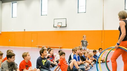 In a sports hall, a group of students sit on the floor and look at the standing teacher who is holding several hula hoops in her hand. In the background you can see an orange wall.