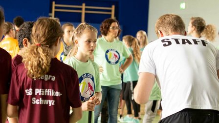 A scene in a gym. A sports teacher with a white T-shirt showing "Staff" is standing in front of a group of students.