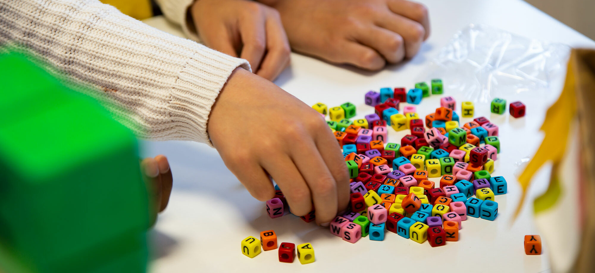 Close-up of three children's hands on a table. One hand reaches into a small pile of colourful letter beads.